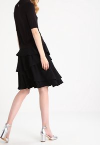 Svart lager-på-lager klänning med en figurnära bodice och volangskikt. Matchad med silverfärgade klackskor med ankelslinga. Slät tyg och strukturerad silhuett.