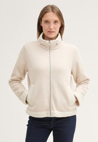 Veste zippée beige clair avec un col haut, doublure en polaire douce, poches latérales et bordures dures aux poignets et à l'ourlet.