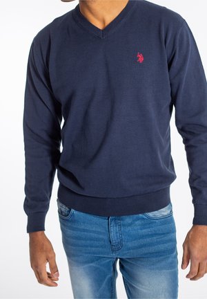 Navyblauw v-halstrui van gebreide stof, met een rood logo op de borst en geribde manchetten en zoom. Draagt over lichtblauwe jeans.