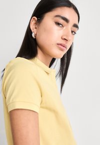 Žluté polo tričko s límcem, krátkými rukávy a knoflíkovou légou. Model má rovné tmavé vlasy a kovové kruhové náušnice.