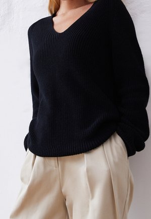 Femme portant un pull noir en maille côtelée à col en V et un pantalon plissé beige, les mains dans les poches, sur un fond clair.