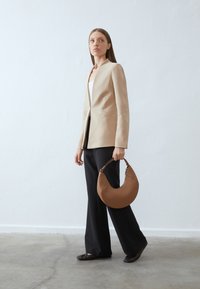 Casaco bege alinhado com design estruturado, combinado com calças pretas. Bolsa em forma de crescente, castanha escura, com uma alça texturizada.
