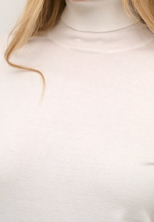 Kvinde iført en lys beige rullekravesweater med langt blondt hår, der falder over hendes skulder mod en ensfarvet baggrund.