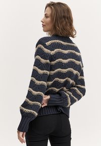 Gebreide trui in marineblauw met beige golvende strepen. Heeft een hoge hals, geribde manchetten en onderkant. De gestructureerde, chunky draad zorgt voor diepte.
