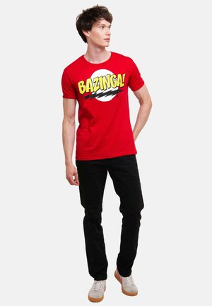 Red cotton t-shirt featuring bold yellow "BAZINGA!" text with black accents, paired with black pants and grey sneakers.