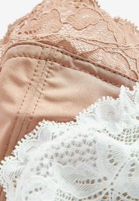 Nackter Satin-BH mit weißer Spitzenbordüre, der florale Muster und gewellte Kanten aufweist. Glatte Textur mit definierten Nähten und Stichen.