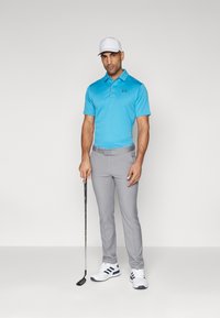 Camiseta polo de color azul claro, pantalones grises, gorra blanca, sosteniendo un palo de golf. Presenta un logo en contraste en la camiseta. Los zapatos son blancos con rayas azules.