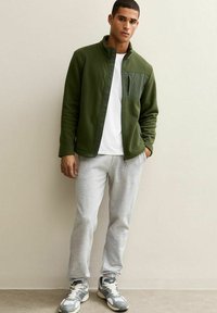 Chaqueta de forro polar verde con cierre de cremallera y bolsillo frontal, combinada con pantalones de chándal gris claro y zapatillas deportivas con detalles blancos.