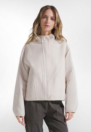Mujer con cabello rubio largo que lleva una chaqueta acolchada beige claro y pantalones oscuros sobre un fondo blanco liso.