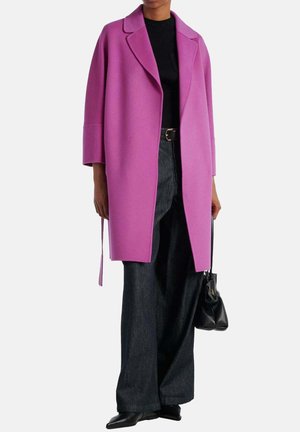 Vrouw draagt een felpaarse oversized jas, een zwart topje, wijde donkere jeans, zwarte schoenen en houdt een kleine zwarte handtas vast.