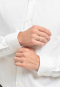 Silver vigselring på en hand som justerar manschetten på en vit skjorta, vilket framhäver den släta texturen och de klassiska knappdetaljerna.