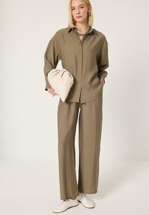 Kobieta w luźnej oliwkowo-zielonej koszuli zapinanej na guziki i pasujących spodniach, trzyma beżową kopertówkę, ma białe trampki i krótkie blond włosy.