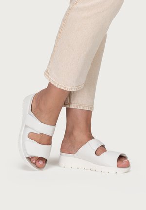 Feet wearing white open-toe sandals with thick soles and adjustable straps, paired with light beige cropped pants against a plain background.