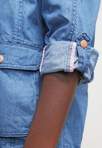 Denimjacka med ärm med uppvikt manschett; ljusblått tyg; randig foder i rosa och vitt; knappelement vid manschetten.