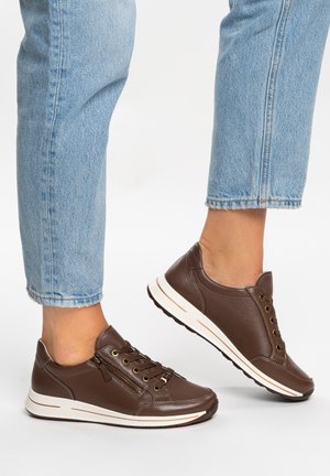 Braune Ledersneaker mit weißen Sohlen, seitlichem Reißverschluss und Schnürsenkeln, getragen zu hellblauen, kurz geschnittenen Jeans vor einfachem Hintergrund.