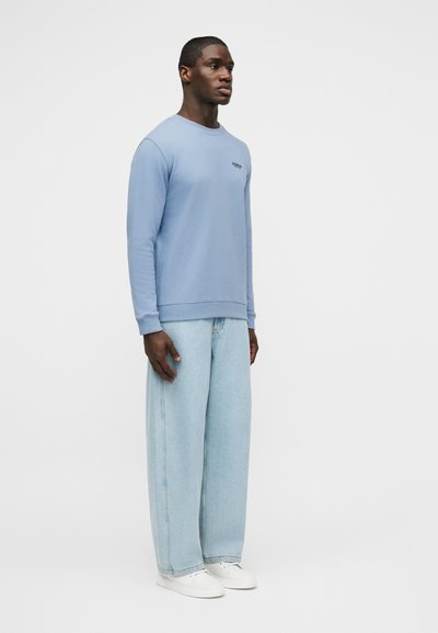 Άνδρας που φοράει ανοιχτό μπλε φούτερ, φαρδιά τζιν ανοιχτής πλύσης και λευκά αθλητικά παπούτσια, στέκεται μπροστά σε απλό γκρίζο φόντο.