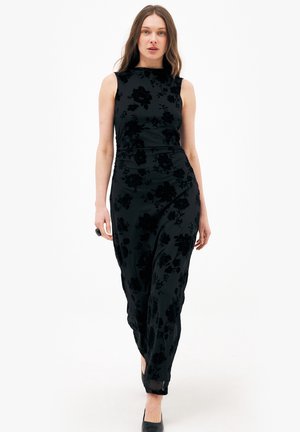 Mujer caminando hacia adelante con un vestido maxi negro sin mangas con un patrón floral de terciopelo y tacones negros sobre un fondo blanco.