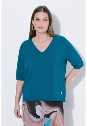 Kvinde iført teal V-hals kortærmet top, store gyldne øreringe i ringform og flerfarvede bukser med abstrakt mønster, stående foran en hvid baggrund.