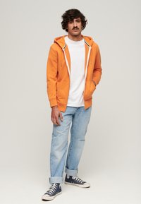 Orange zip-up hoodie med hvide snore, hvid t-shirt med rund hals under, lyseblå jeans og navy canvas sneakers.