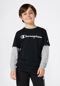 Juodas trumpas rankovėmis marškinėliai su baltu "Champion" logotipu, uždėti ant pilkų ilgomis rankovėmis marškinėlių. Suderinti su juodomis kelnėmis, kurios turi logotipo detalę.