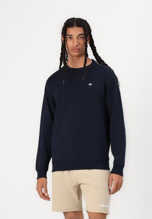 Marineblå sweater i et blødt, struktureret stof med lange ærmer, rund halsudskæring og et lille logo på brystet. Kombineret med beige shorts.