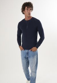 Granatowa, prążkowana koszula henley z długim rękawem i trzema guzikami, w połączeniu z jasnoniebieskimi dżinsami typu straight-leg oraz białymi sneakersami.