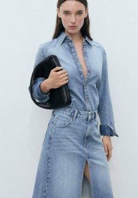 Denim srajca z ovratnikom na gumb, svetlo modre barve. V kombinaciji z usklajeno denim minico z sprednjim razporkom. Drži teksturirano črno torbico.