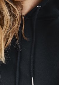 Svart huvtröja med dragkedja som har silverdetaljer och en mjuk, texturerad väv. Nära vy som framhäver halsringningen och tygets detaljer.