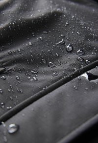 Tessuto nero, testurizzato con gocce d'acqua. Inquadratura ravvicinata che mostra una superficie liscia e una fine trama, evidenziando la resistenza all'acqua.