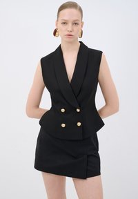 Schwarzer ärmelloser Blazer mit tiefem V-Ausschnitt, doppelt geknöpften goldenen Akzenten, tailliertem Schnitt, kombiniert mit einem passenden schwarzen Minirock.