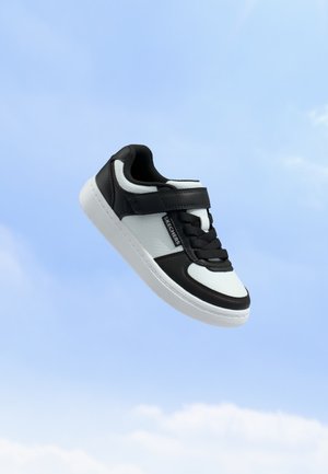 Scarpa Skechers bianca e nera con lacci e cinturino in velcro che fluttua su uno sfondo di cielo azzurro limpido.