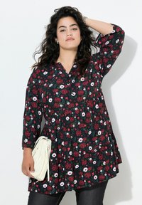 Blumen-Tunika mit tiefem V-Ausschnitt und gefälteltem Design, verziert mit roten und weißen Blumen auf schwarzem Hintergrund. Lange Ärmel, leichtes Material, kombiniert mit einer weißen Tasche.