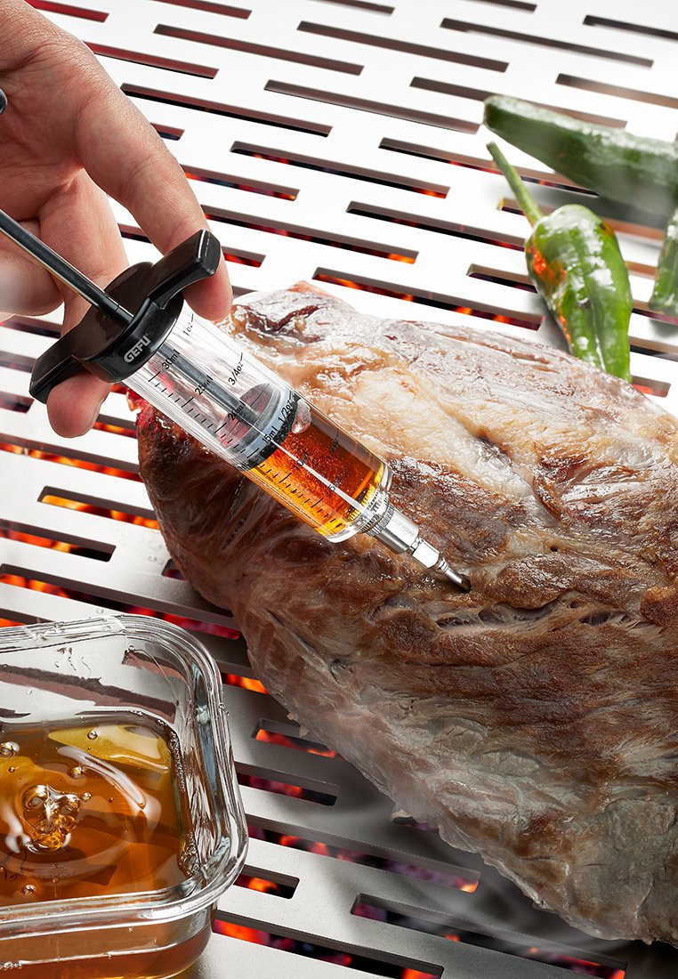 Injektionsspritze mit klaarem Kunststoffkörper und schwarzen Akzenten injiziert Marinade in ein gebräuntes, strukturiertes Stück Fleisch auf einer Metallgrilloberfläche.