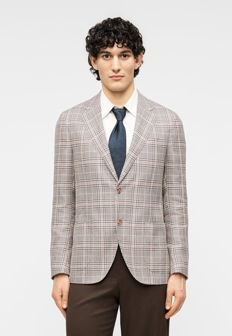 Ανοιχτό γκρι καρό σακάκι με καφέ λεπτομέρειες, κούμπωμα με ένα κουμπί, γιακάδες με σχισμές, φορεμένο πάνω από λευκό πουκάμισο και σκούρα γραβάτα, υφασμάτινο μείγμα.