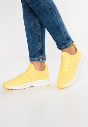 Osoba w niebieskich dżinsach i żółtych siateczkowych trampkach z białymi podeszwami stoi na jasnej podłodze na tle białej ściany.