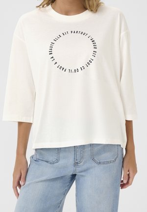 Vrouw draagt een wit shirt met lange mouwen met Franse tekst in een cirkel en lichtblauwe jeans, staand tegen een effen achtergrond.