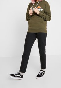 Olivgrön huvtröja med en vit logotyp, kombinerad med svarta jeans och svarta sneakers med vita detaljer. Justerbara dragsnören vid huvan.