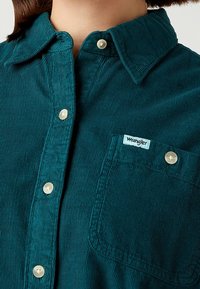 Camicia in corduroy verde blu con colletto button-down, tasca frontale e bottoni bianchi. Presenta un'etichetta "Wrangler" sulla tasca.