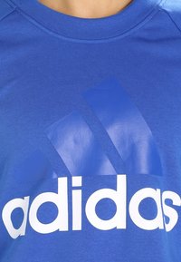 Camiseta de algod�ón azul con cuello redondo que presenta un gran logo blanco de Adidas y tres rayas diagonales azules en la parte superior.