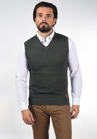 Mørkegrøn strikvest med V-hals design. Textureret stof, ribbet bundkant, båret over en hvid skjorte, kombineret med brune bukser.