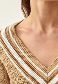 Nahaufnahme des Halses und oberen Brustbereichs einer Person, die einen beigen Strickpullover mit einem braun-weißen gestreiften V-Ausschnitt trägt.