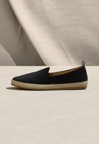 Chaussure espadrille noire à enfiler avec semelle en jute beige tissée et languette au talon, posée sur une surface claire devant un fond en tissu.