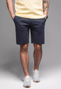 Pantaloni corti grigi in cotone con una vestibilità rilassata, dotati di una fascia elastica in vita e tasche laterali, abbinati a scarpe da ginnastica grigio chiaro.