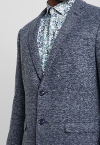 Marinblå strukturerad kavaj med notch lapel, två knappar och en ficka; bärs över en mönstrad skjorta med knappar i gröna och blå nyanser.