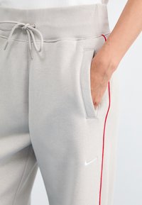 Sive hlače s vrvico v pasu, rdečimi stranskimi črtami in belim Nike logotipom na levem stegnu. Mehka tkanina in stranski žepi.