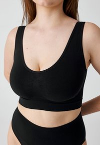 Frau trägt einen schwarzen nahtlosen Sport-BH und hoch taillierte Unterwäsche, beschnitten vom Hals bis zu den Hüften, steht vor einem neutralen Hintergrund.