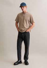 Beżowy bawełniany t-shirt z małym napisem, do tego czarne dżinsy i czarne buty. Stylizację dopełnia ciemnoniebieska czapka.