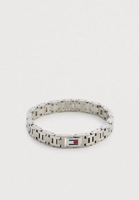 Zilverkleurige metalen armband met in elkaar grijpende schakels, voorzien van een rechthoekige sluiting met een rood-blauw logo. Gladde, gepolijste textuur.