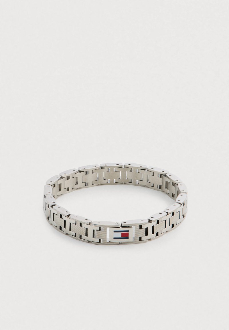Zilverkleurige metalen armband met in elkaar grijpende schakels, voorzien van een rechthoekige sluiting met een rood-blauw logo. Gladde, gepolijste textuur.
