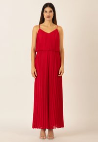 Rotes, plissiertes Maxikleid mit dünnen Trägern und einem geschichteten Design, das eine glatte Textur und eine fließende Silhouette bietet. Silbergraue Riemchensandalen.